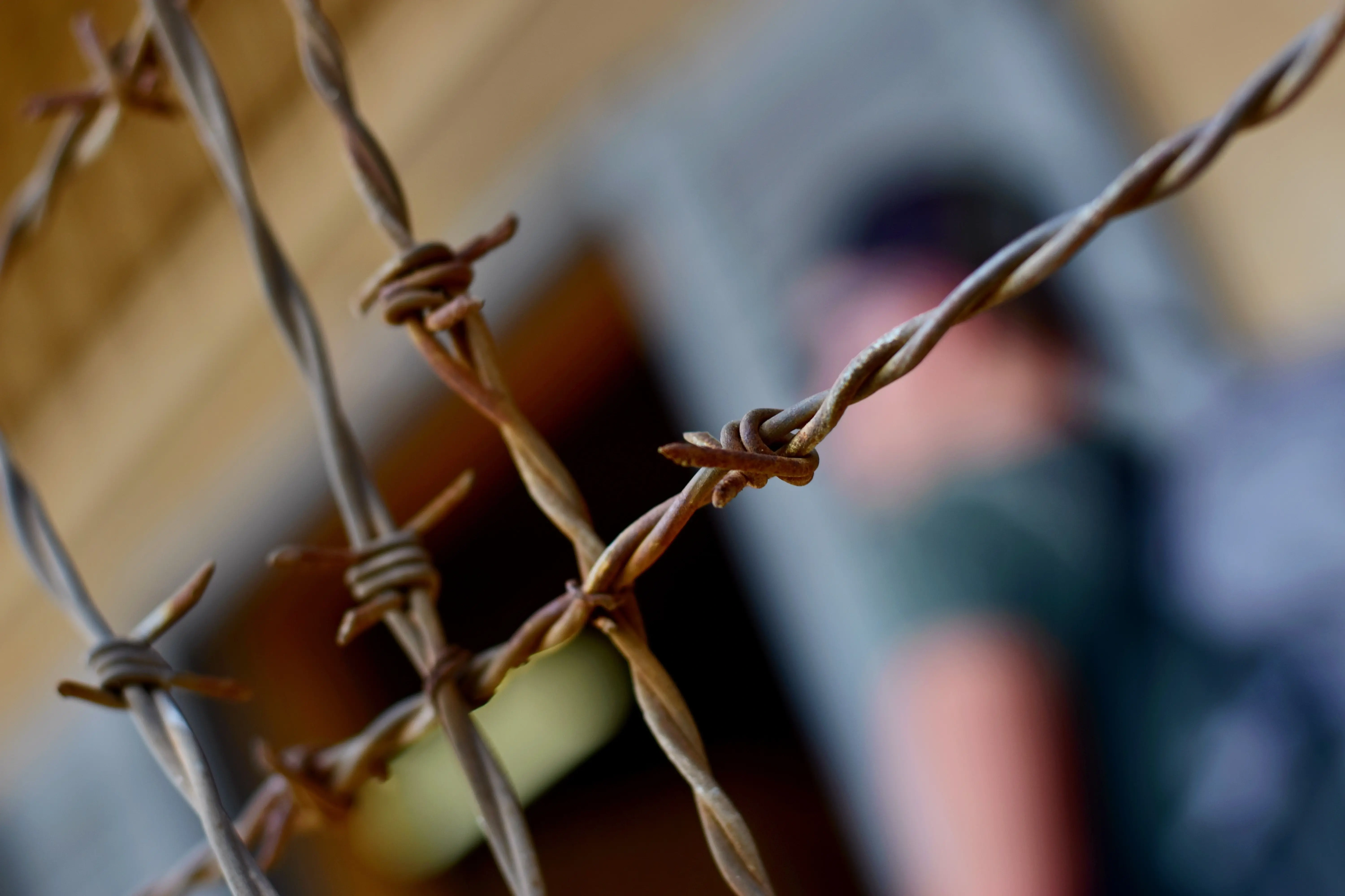 Detail rezavého ostnatého drátu v popředí s rozmazaným pozadím, které naznačuje přítomnost osoby. Ostnatý drát symbolizuje překážky a hranice.